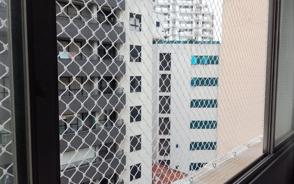 Rede de proteção branca em janela de apartamento, vista de dentro, com prédios ao fundo